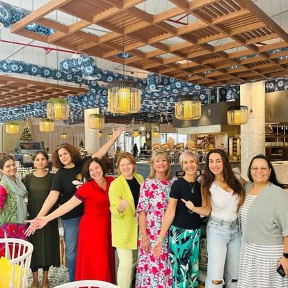 A wide-angle shot of nine women posing together in a brightly lit, modern restaurant. They are dressed in a variety of colorful casual and smart-casual attire, including a bright red dress, a lime green blazer, and a floral pink dress. The restaurant features a unique wooden trellis ceiling with decorative blue patterned ductwork and hanging cylindrical pendant lights.
