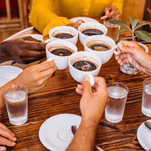 An overhead, close-up shot of a diverse group of people bringing five white ceramic mugs of black coffee together for a toast over a rustic wooden table. Several glasses of iced water, white saucers, and a small potted succulent are scattered across the table. One person in the background wears a bright yellow long-sleeved shirt, and the scene is filled with warm, natural light.