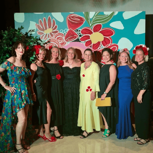 A group of eight women posing together in front of a colorful, floral-themed backdrop at an indoor event. The women are dressed in elegant evening wear, including long gowns and cocktail dresses in colors like black, royal blue, yellow, and a floral print. Several women are wearing decorative flower crowns or floral hair accessories, and one holds a yellow clutch bag. The lighting is warm and festive, typical of a gala or celebration.