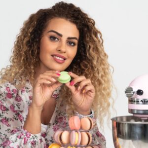 Macaron House Oman baker holding a green macaron with a tiered display of French macarons, homemade macaron business in Muscat